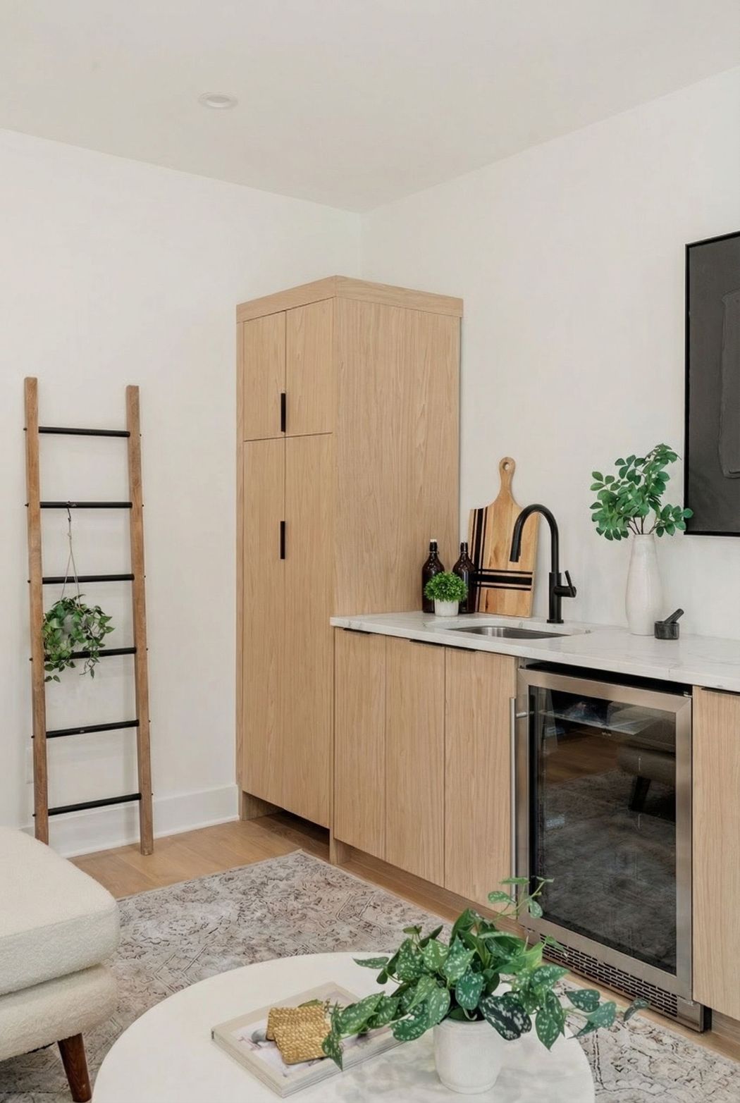 A kitchenette with light wood cabinets, a glass-front beverage cooler, and a decorative wooden ladder against a white wall.