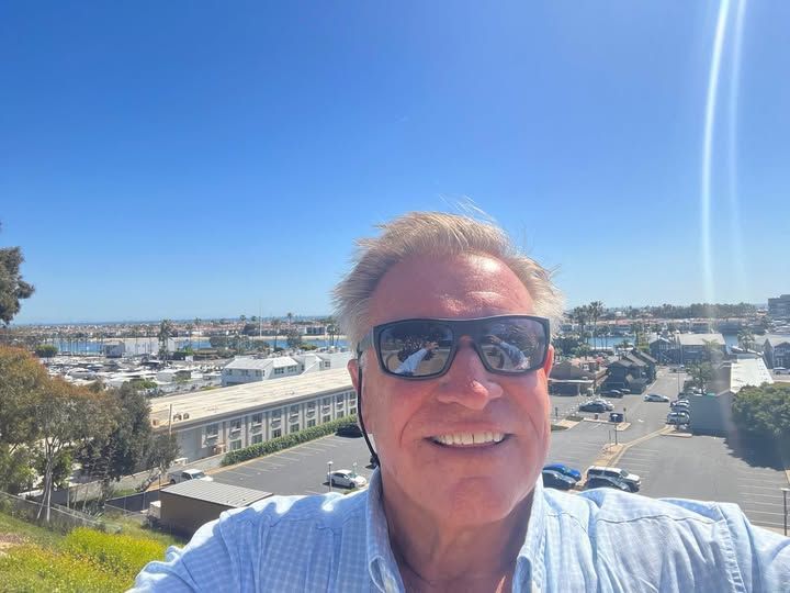 A man wearing sunglasses is taking a selfie with a city in the background.