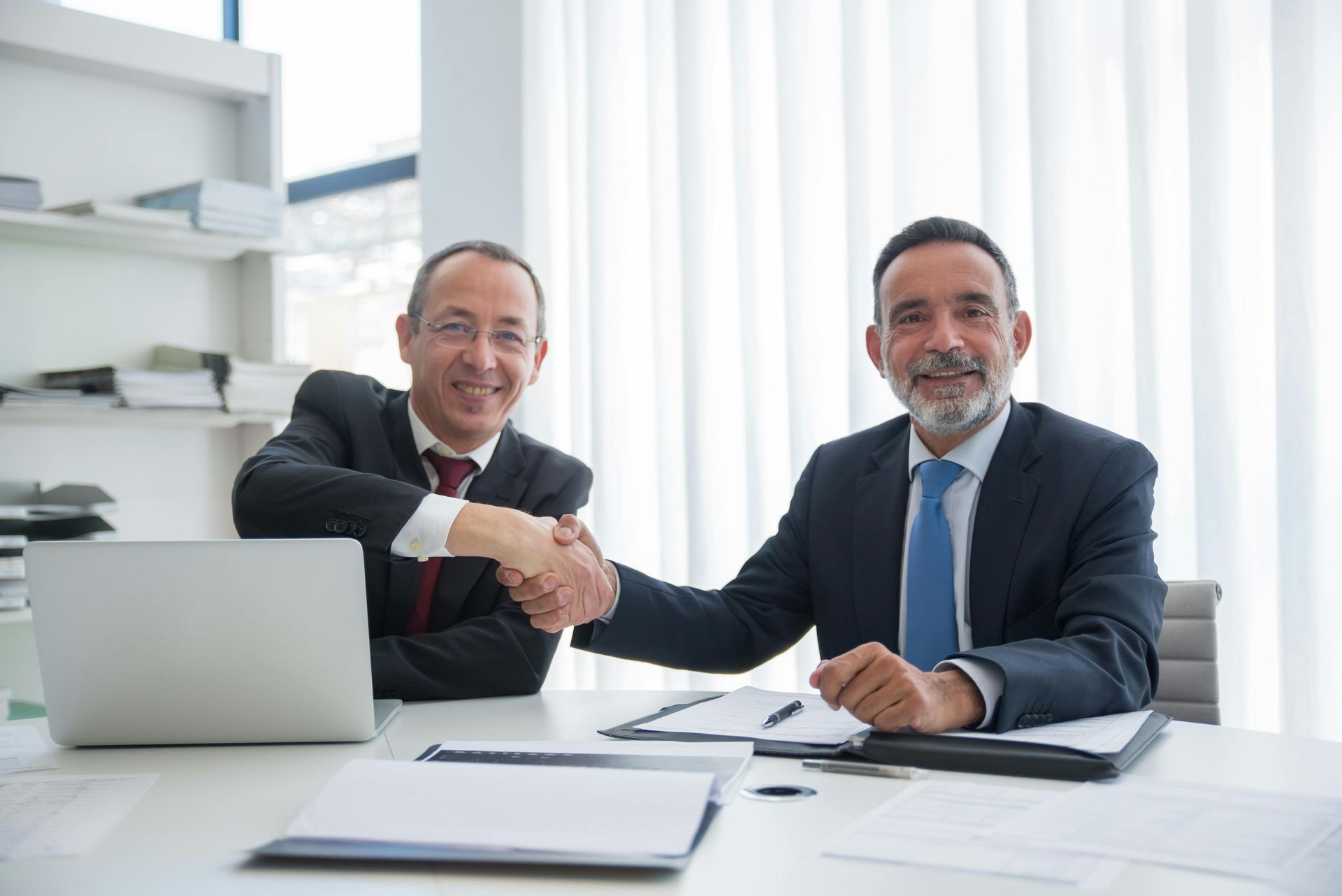 Two businesspeople shaking hands at a desk in a bright office with a laptop and papers