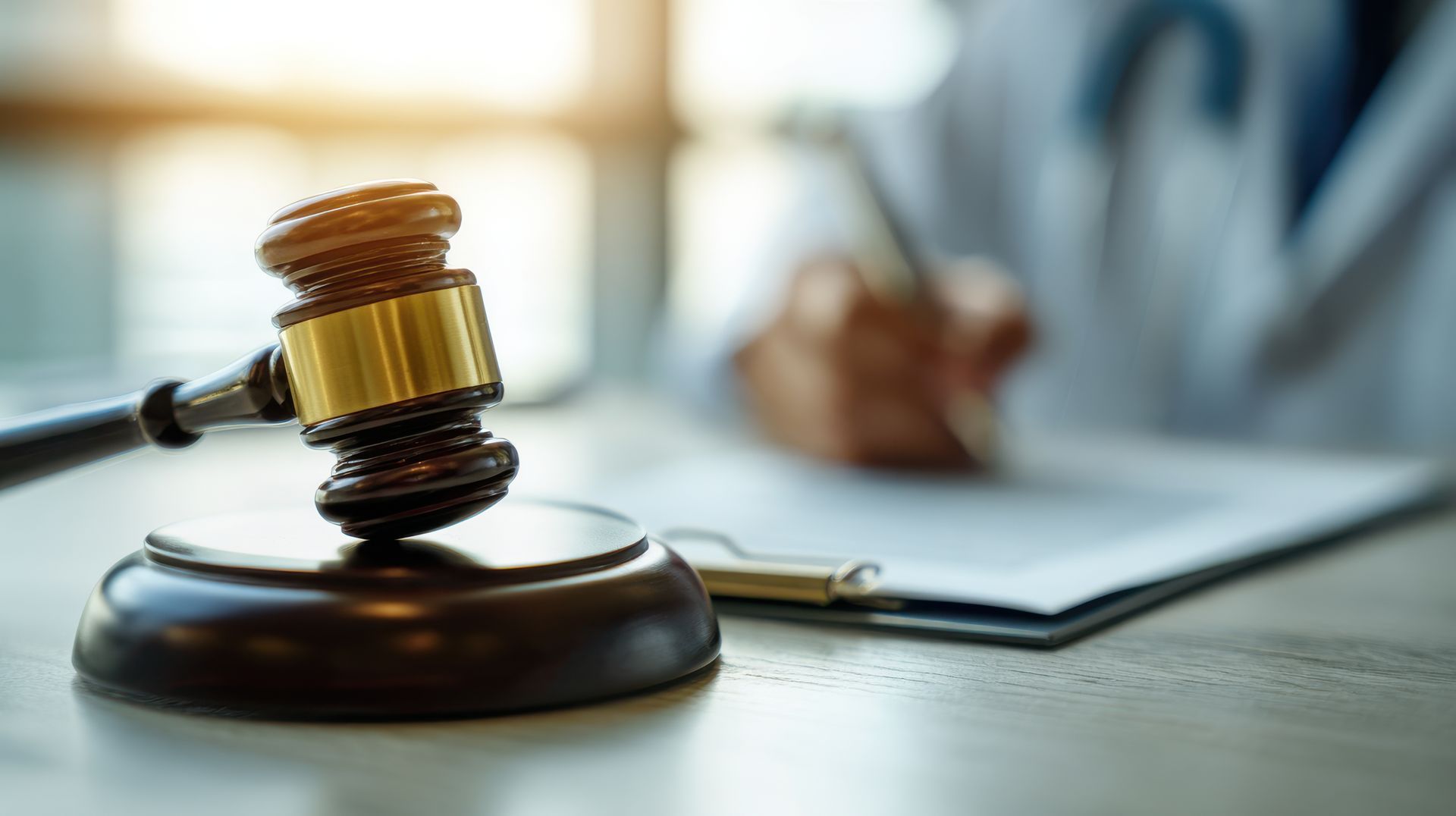 Judge’s gavel on a desk beside a document, with a blurred person writing in the background.