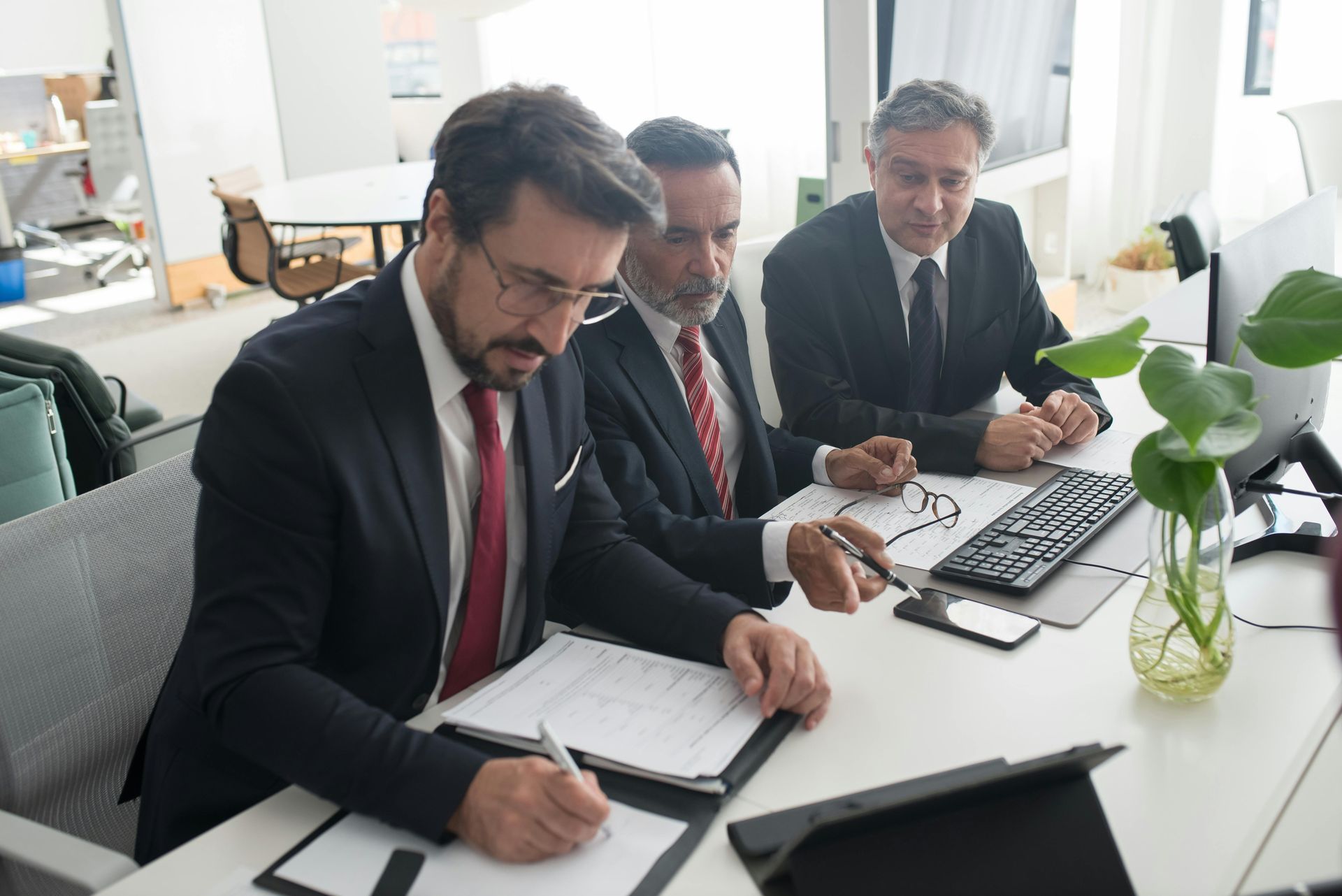 Three men in suits reviewing documents at a desk in a bright office