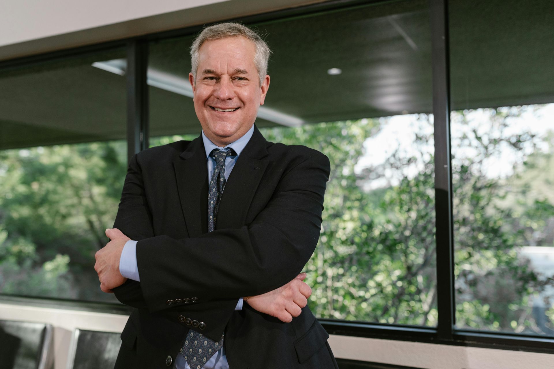 Smiling man in a black suit standing with arms crossed in an office by large windows.