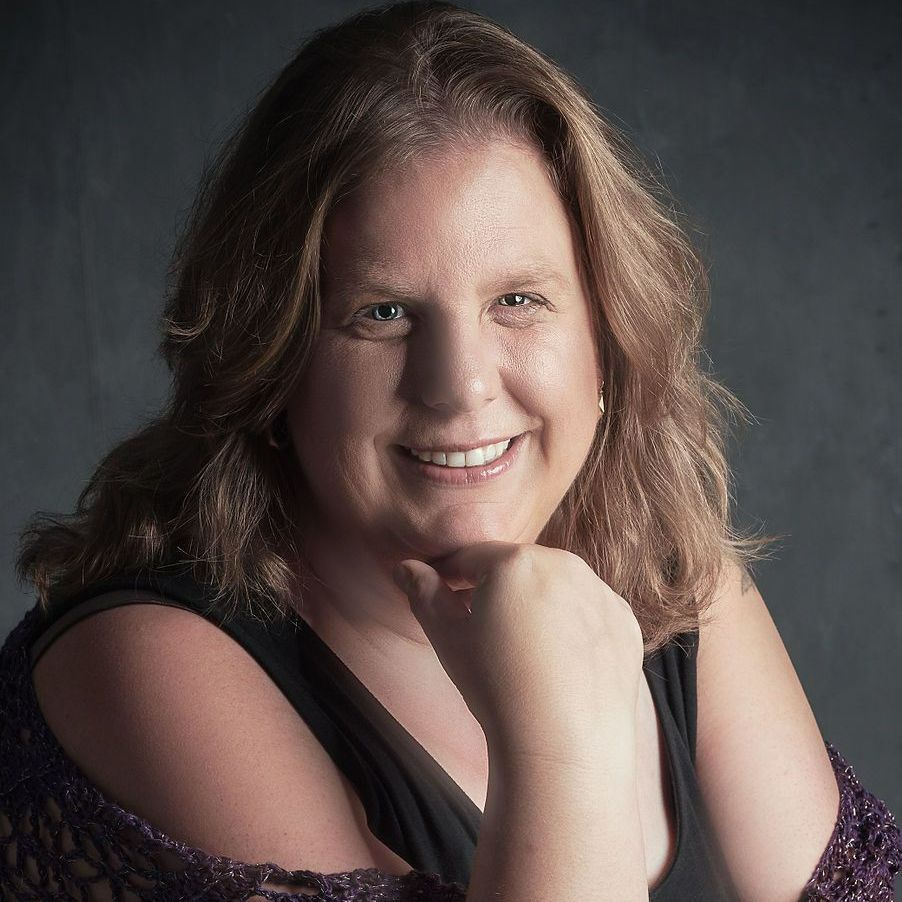 Een vrouw die een paarse omslagdoek en een zwarte top draagt, lacht met haar hand op haar kin.