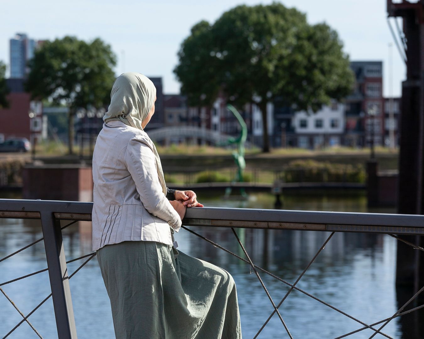 Een persoon met een hoofddoek en lichtgekleurde kleding kijkt op een zonnige dag vanaf een brug uit over een rivier.