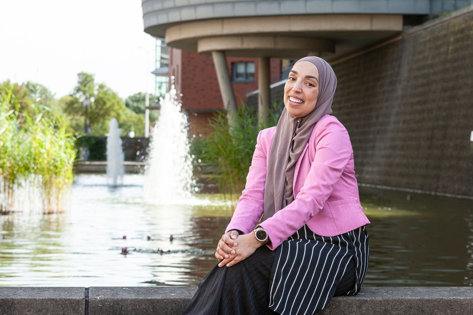 Een lachende persoon, gekleed in een roze blazer en hijab, zit op een richel voor een fontein en een bakstenen gebouw.