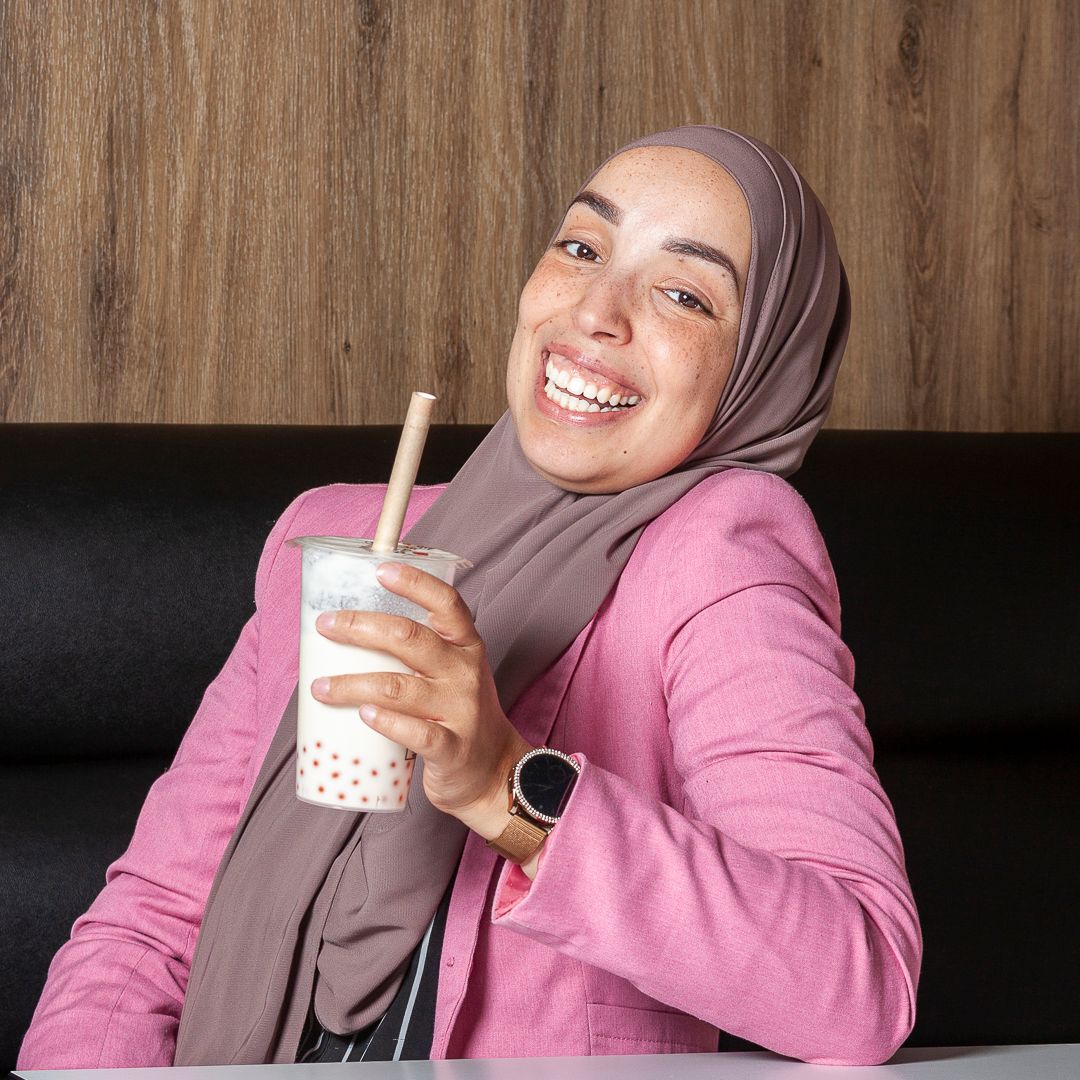 Een persoon met een bruine hijab en een roze blazer glimlacht terwijl ze een kop bubble tea vasthoudt tegen een houten lambrisering.