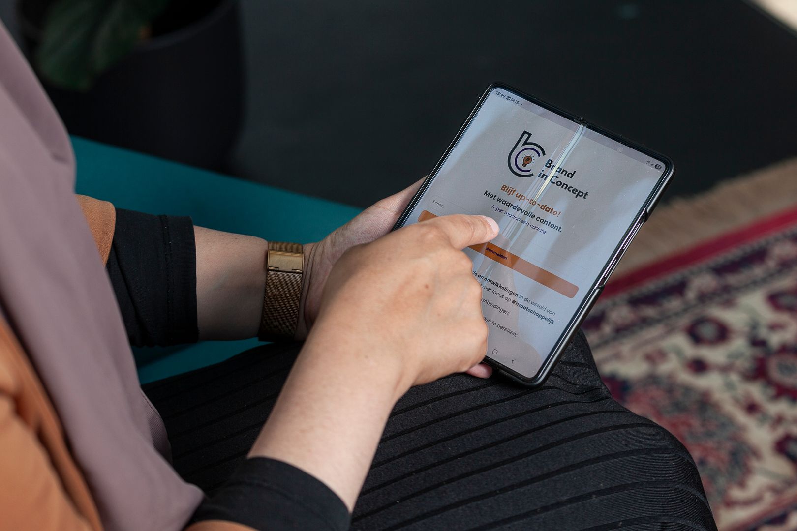 Een persoon met een hijab gebruikt haar vinger om te scrollen op een opvouwbare smartphone terwijl ze op een turquoise bank zit.