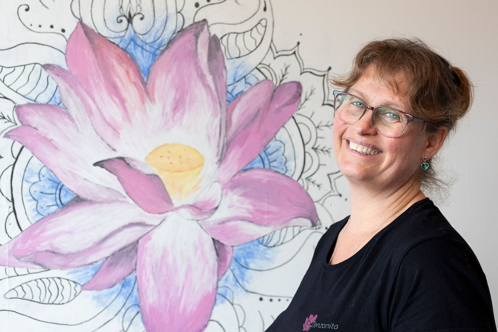 Een persoon lacht naast een grote, kleurrijke muurschildering van een roze lotusbloem tegen een achtergrond met lijntekeningen.
