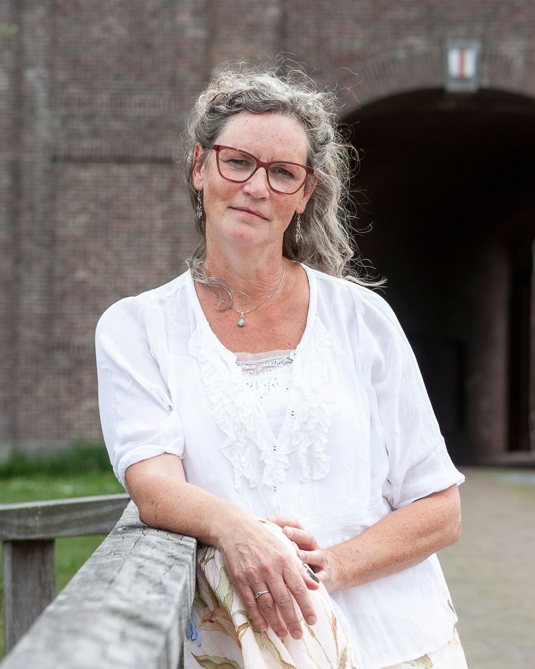 Vrouw met bril, gekleed in een witte blouse en een lichte rok, leunend tegen een houten balustrade, tegen een bakstenen gebouw met een boog.