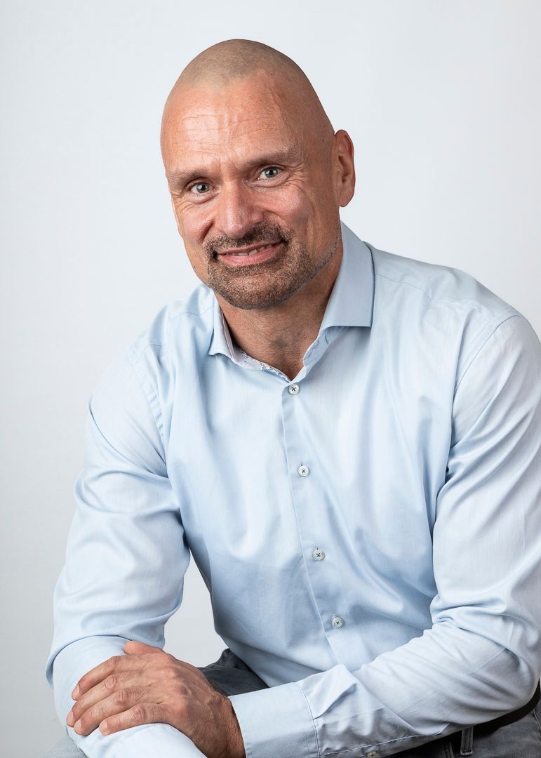 Een kale man met een sikje, gekleed in een lichtblauw overhemd, lacht naar de camera.