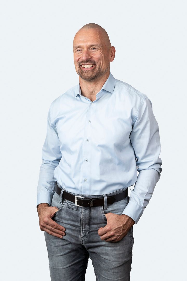 Kale man in een lichtblauw shirt en spijkerbroek, lachend, met zijn handen in zijn zakken, tegen een witte achtergrond.