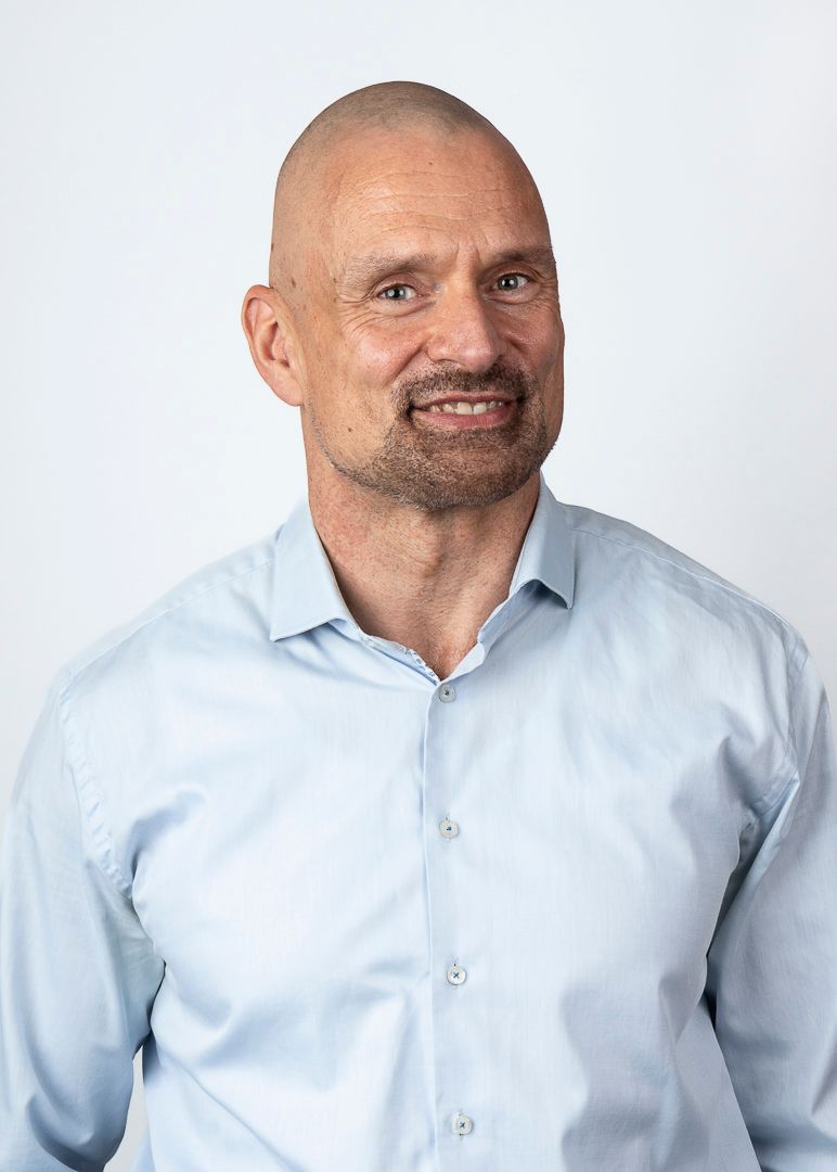 Een kale man met een lichte baard glimlacht, gekleed in een lichtblauw overhemd, tegen een witte achtergrond.