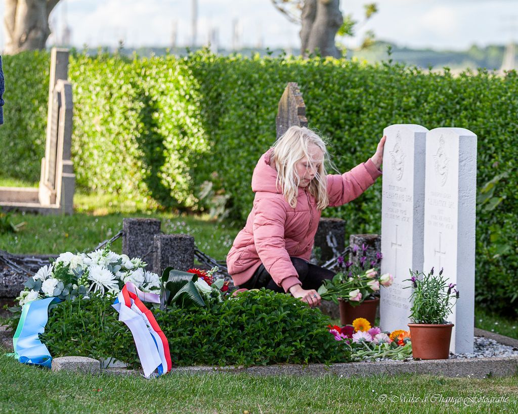 Een persoon knielt bij een graf met een dubbele witte grafsteen en legt bloemen. De omgeving is een groene begraafplaats met een haag.