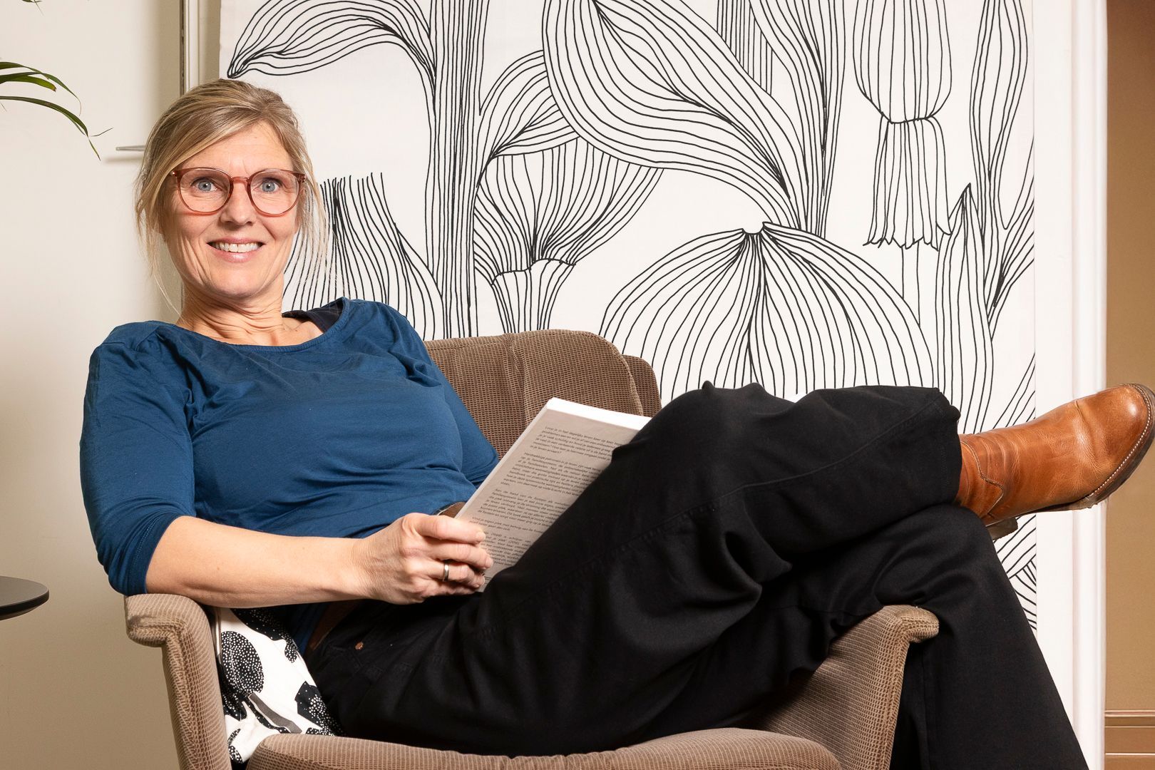 Een glimlachend persoon met een bril zit in een fauteuil en leest een boek voor een behang met een zwart-wit patroon.