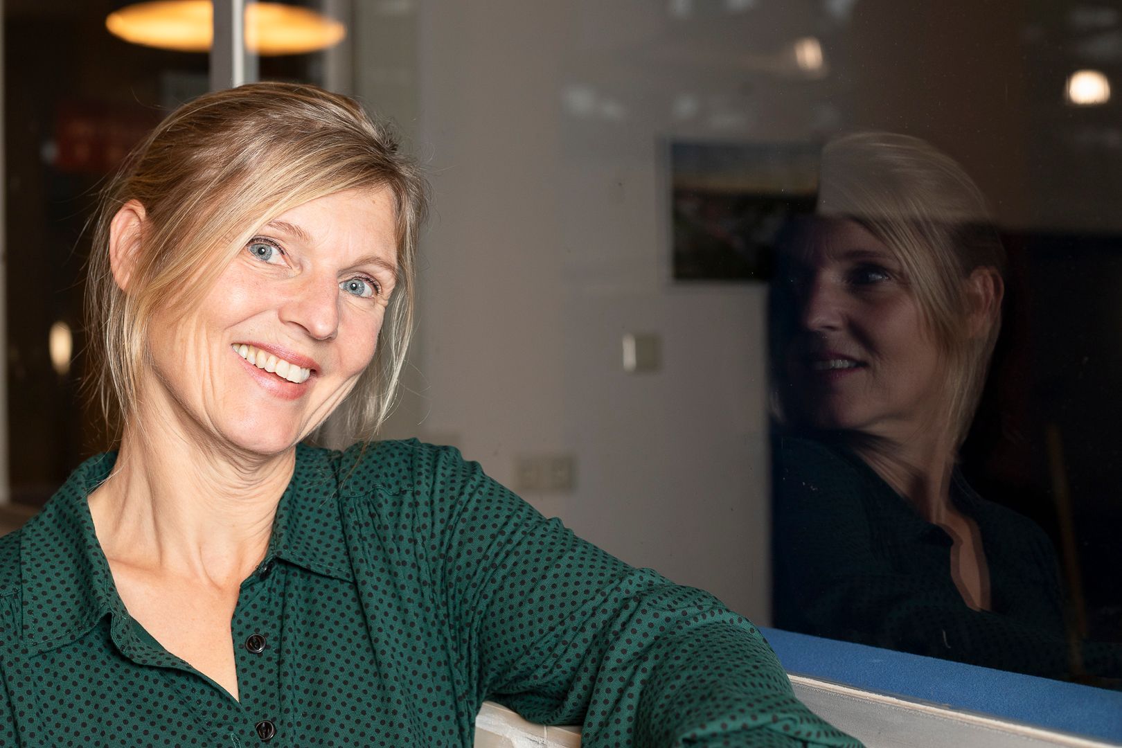 Een lachend persoon in een groen overhemd met patroon, wiens spiegelbeeld zichtbaar is in een raam achter hem of haar.