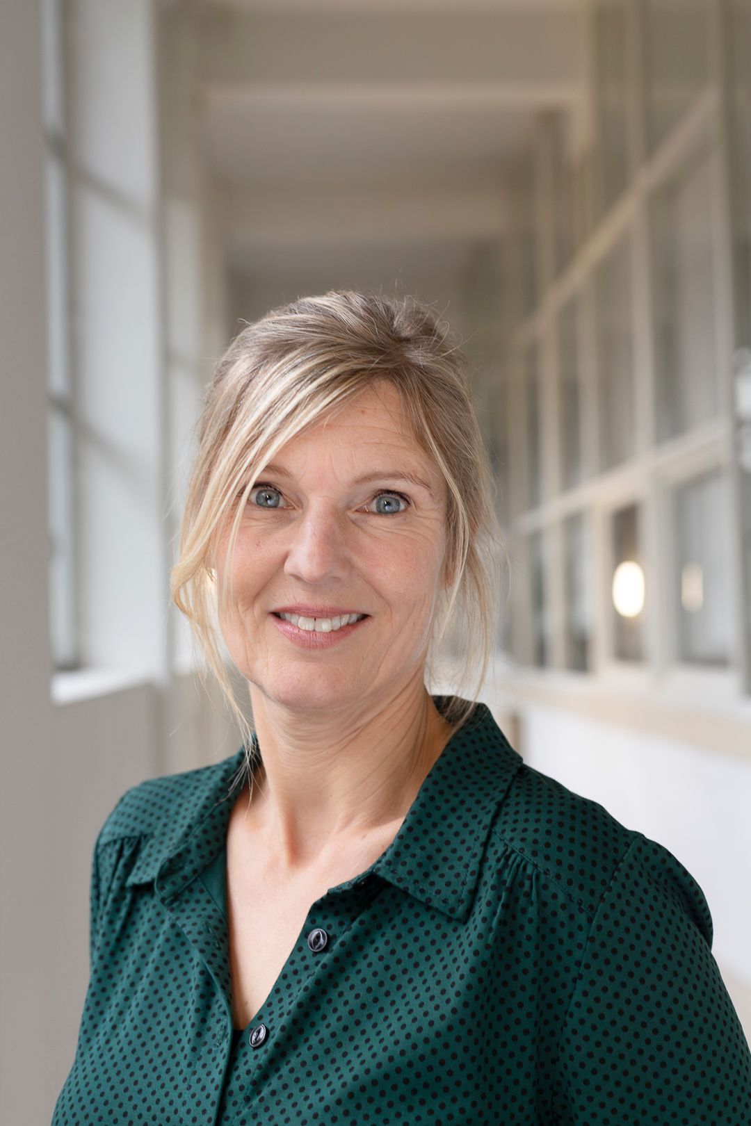 Een glimlachende persoon met blond haar draagt ​​een donkergroen, bedrukt overhemd in een lichte, moderne hal.