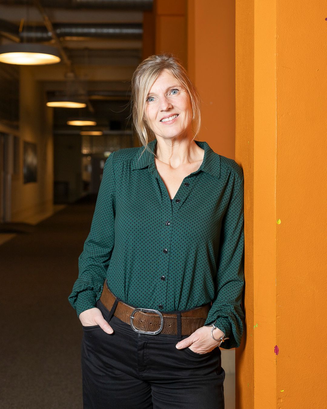 Een persoon, gekleed in een groene blouse met patroon en een zwarte broek met een brede bruine riem, leunt tegen een oranje muur in een gang.