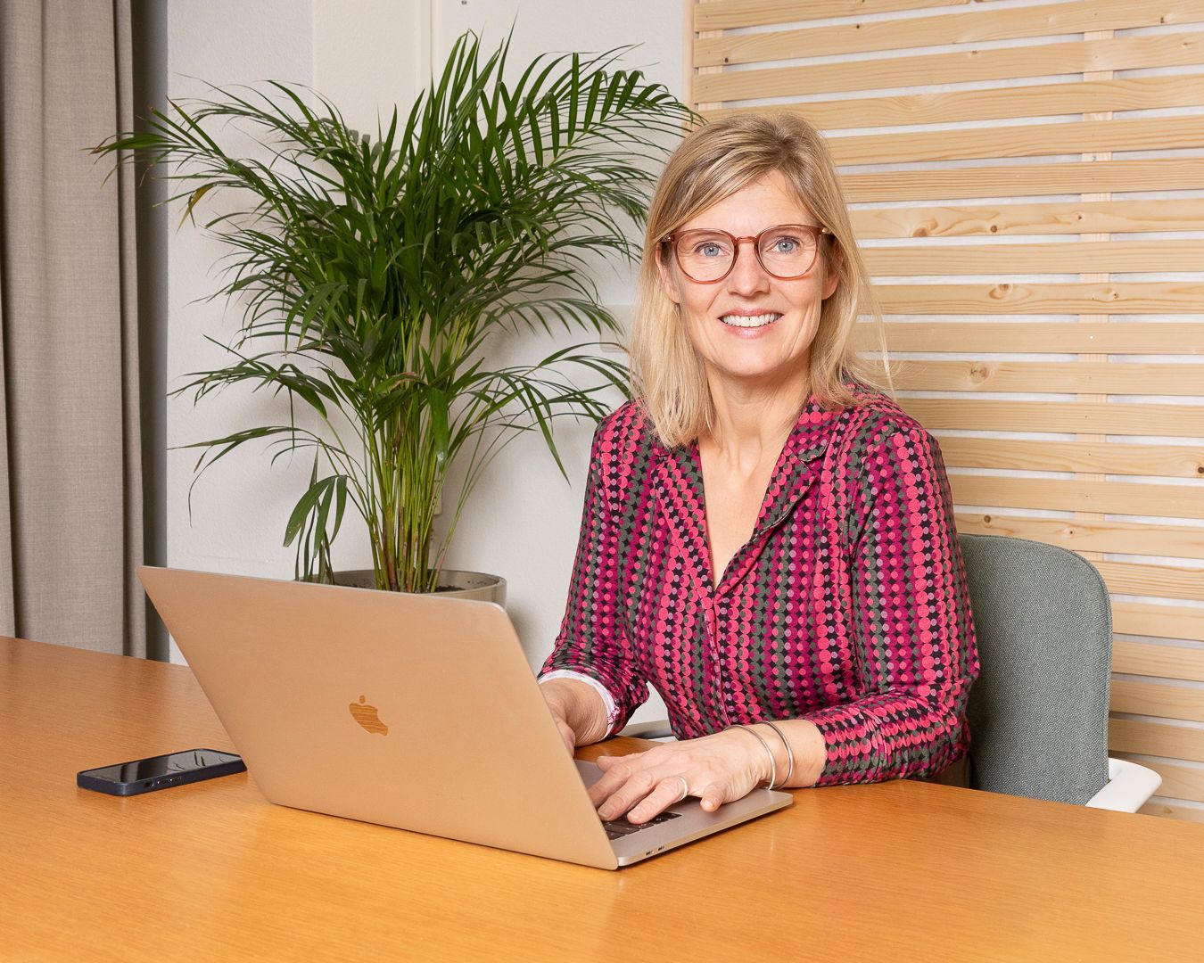 Een vrouw met een bril en een roze blouse met patroon zit aan een bureau te typen op een laptop, met een groene plant op de achtergrond.