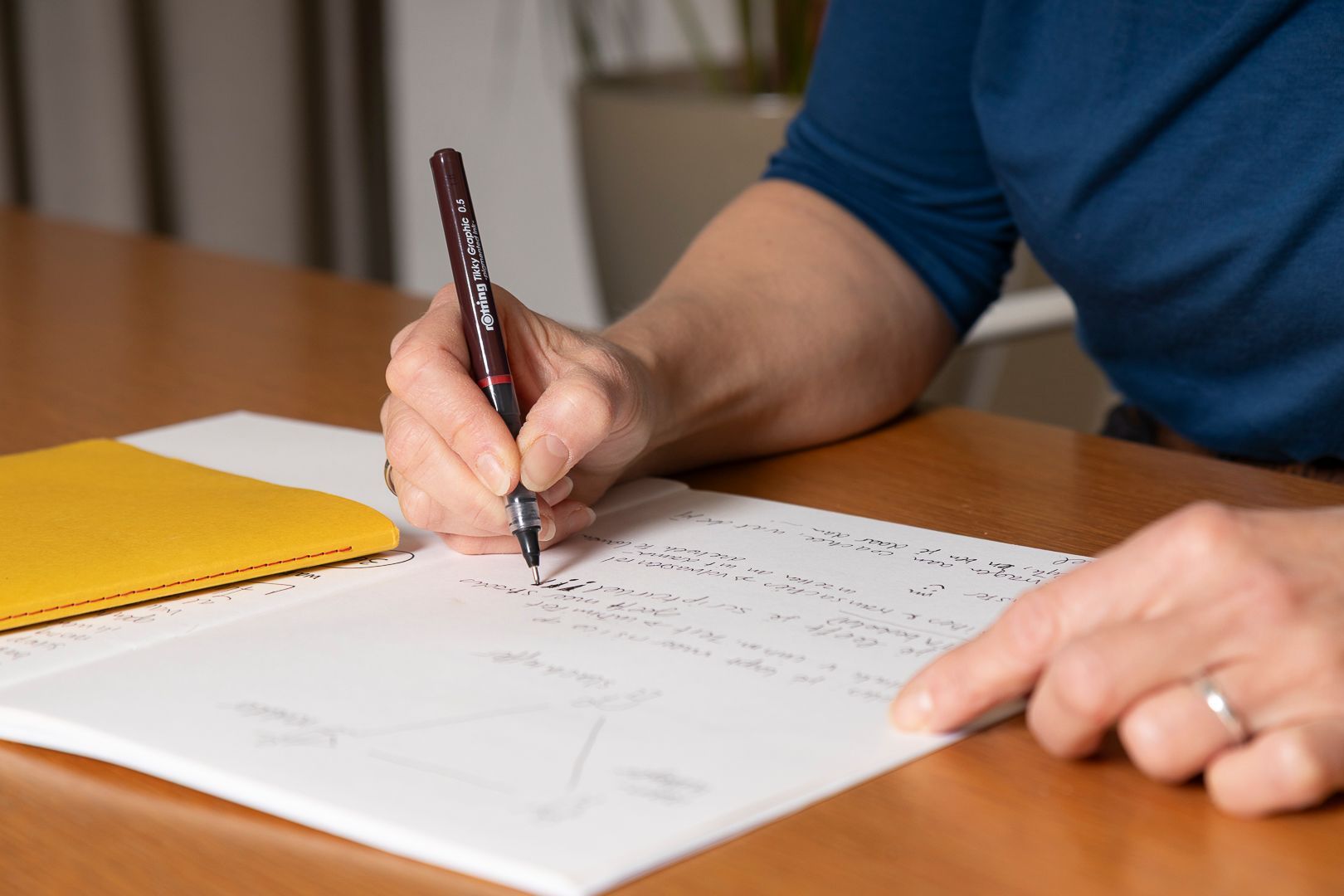Een persoon in een blauw shirt schrijft met een donkere pen op wit papier aan een houten tafel naast een geel notitieboekje.