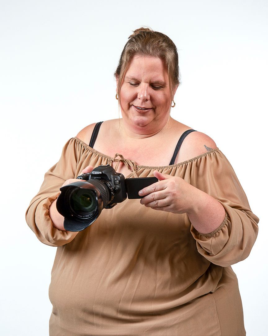 Een vrouw met een digitale camera in haar hand kijkt naar het scherm. Ze draagt een bruine top met blote schouders en een witte achtergrond.