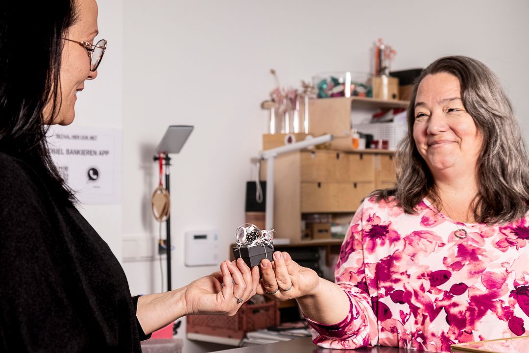 Twee vrouwen wisselen een klein cadeautje uit in een winkel. De ene vrouw glimlacht en overhandigt een ingepakte doos. De andere glimlacht en neemt hem aan.
