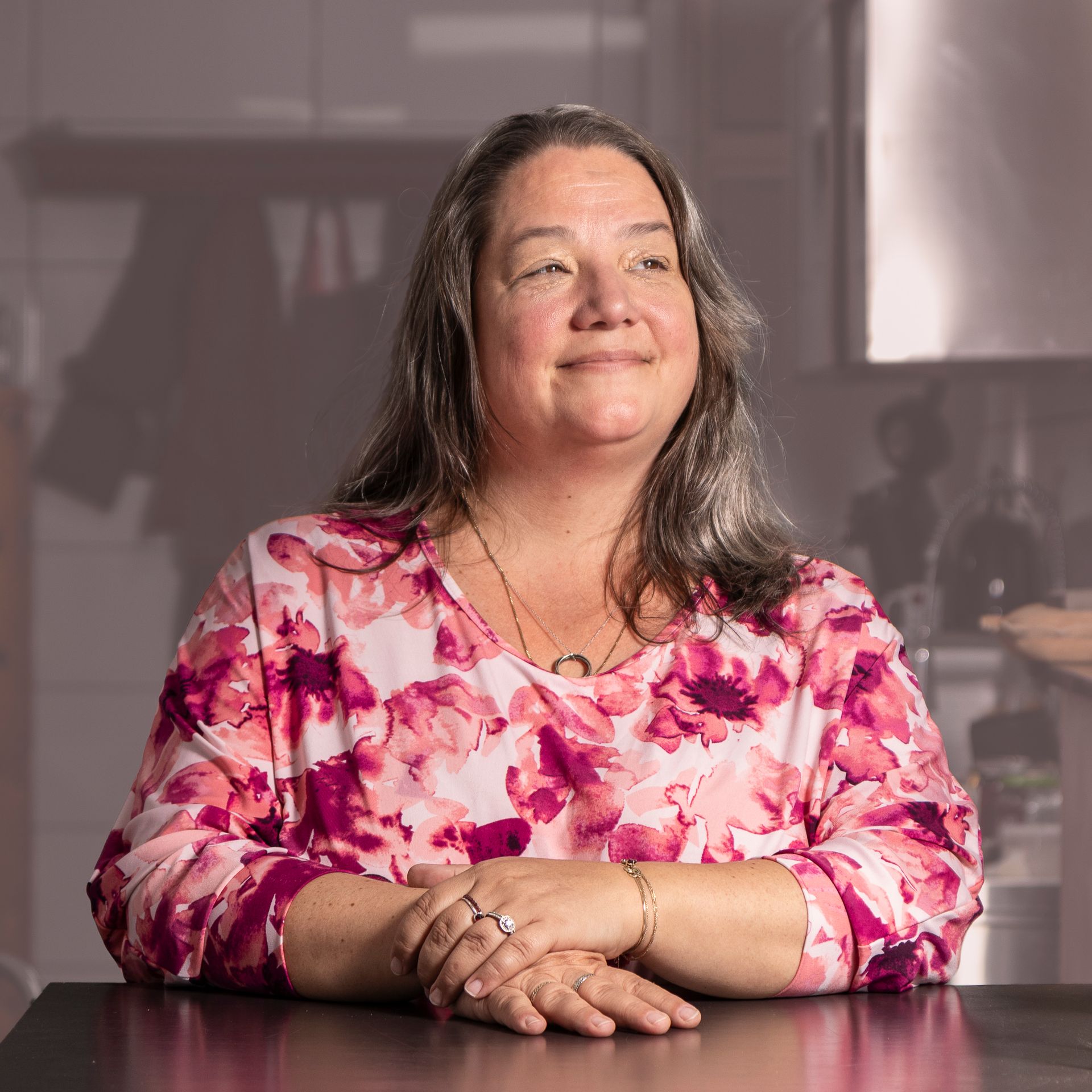 Een vrouw met lang grijs haar en een roze bloemetjesblouse, glimlachend, haar armen rustend op een donkere tafel in wat een keuken lijkt.