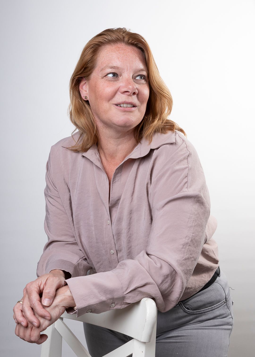 Een vrouw met roodblond haar, gekleed in een lichtpaars shirt en een grijze broek, leunt tegen een witte stoel en kijkt opzij.