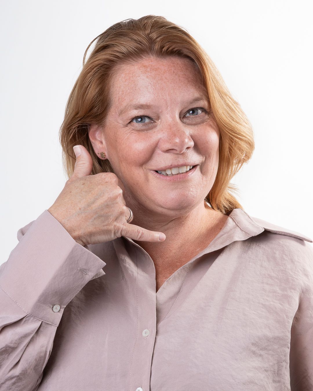 Vrouw met roodblond haar lacht en gebaart met haar hand dat ze moet bellen. Ze draagt een lichtroze overhemd.
