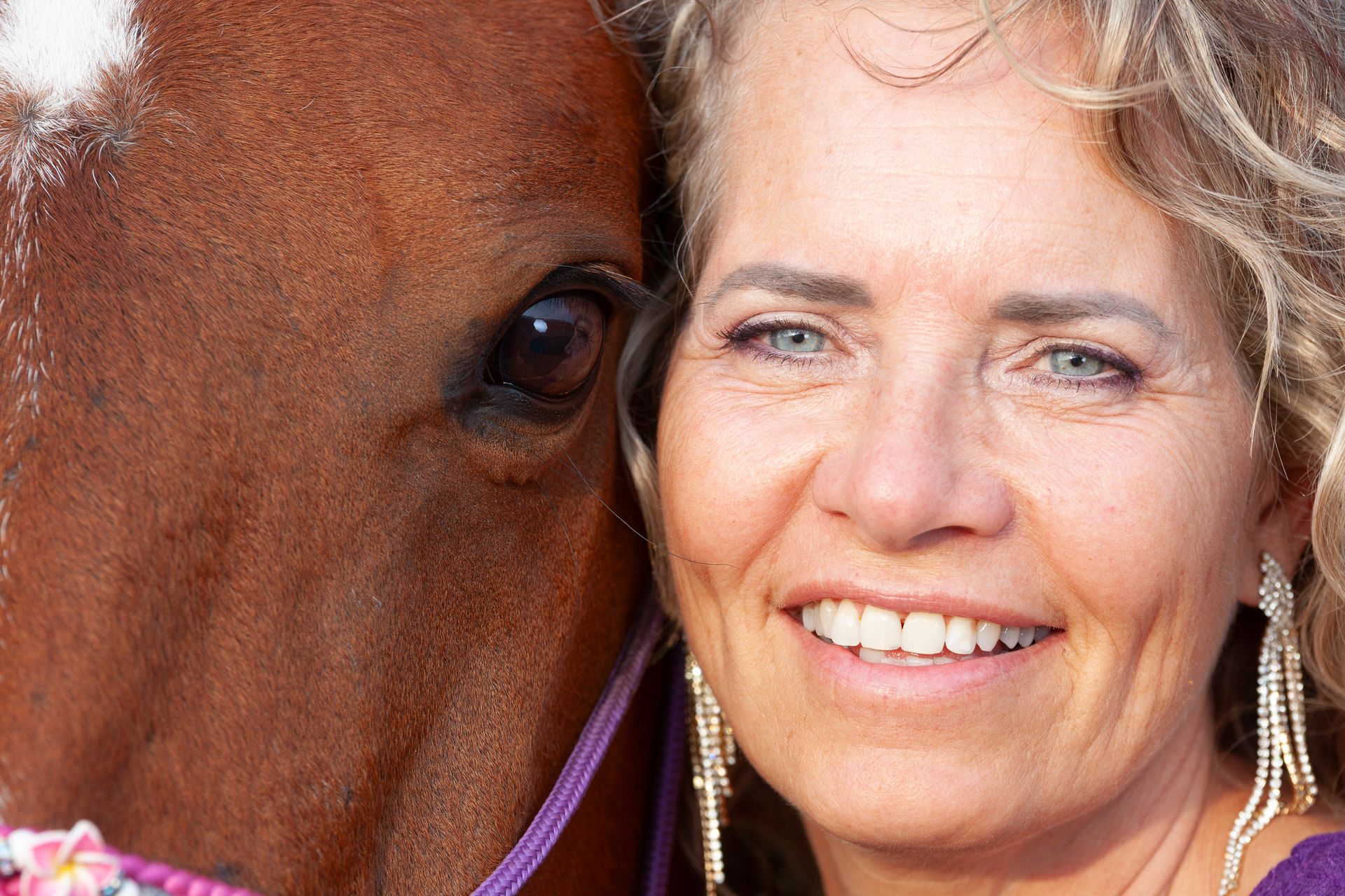 Een lachend persoon met krullend haar poseert dicht naast het hoofd van een paard.