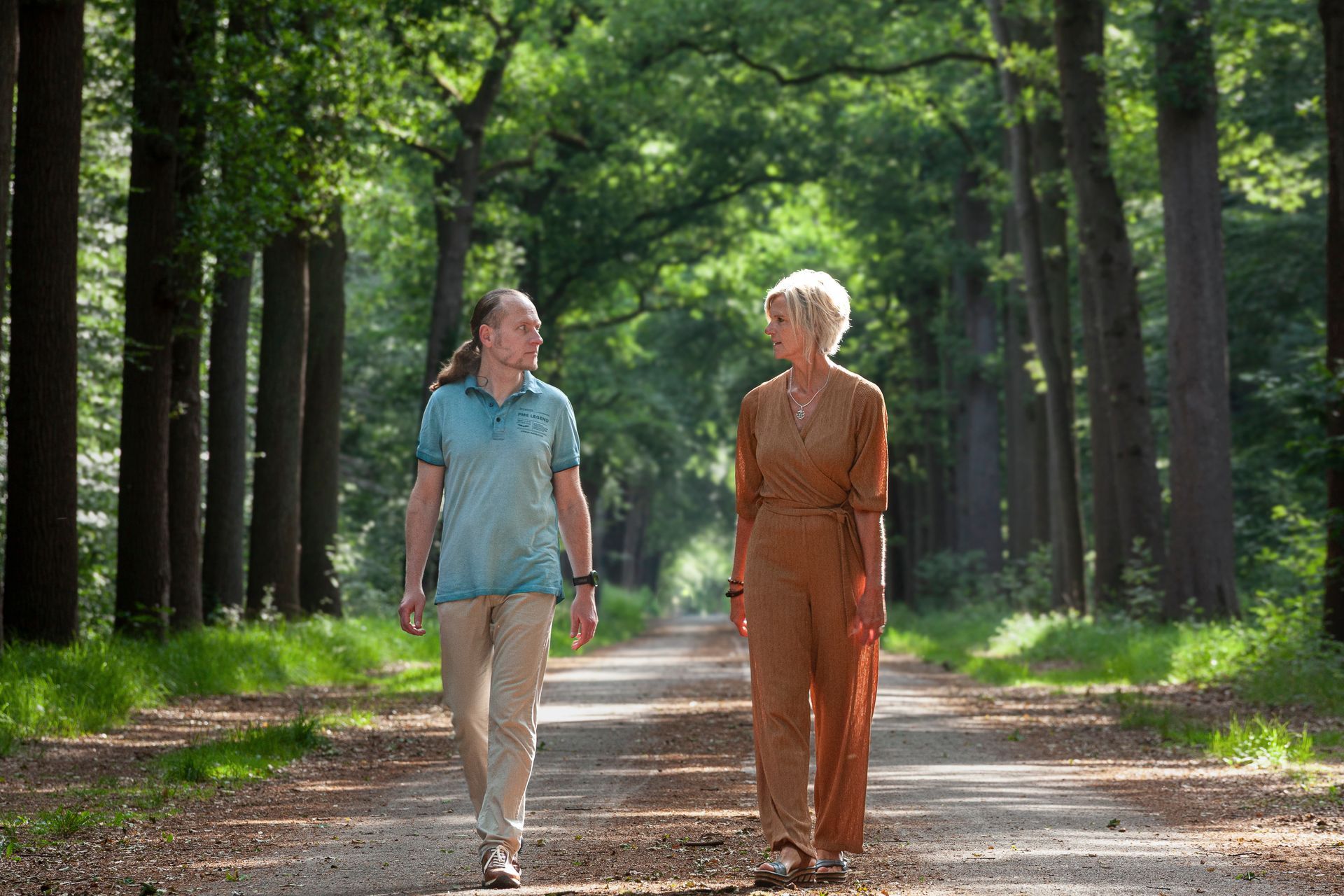 Twee mensen lopen richting de camera over een pad met bomen in een zonnig bos.