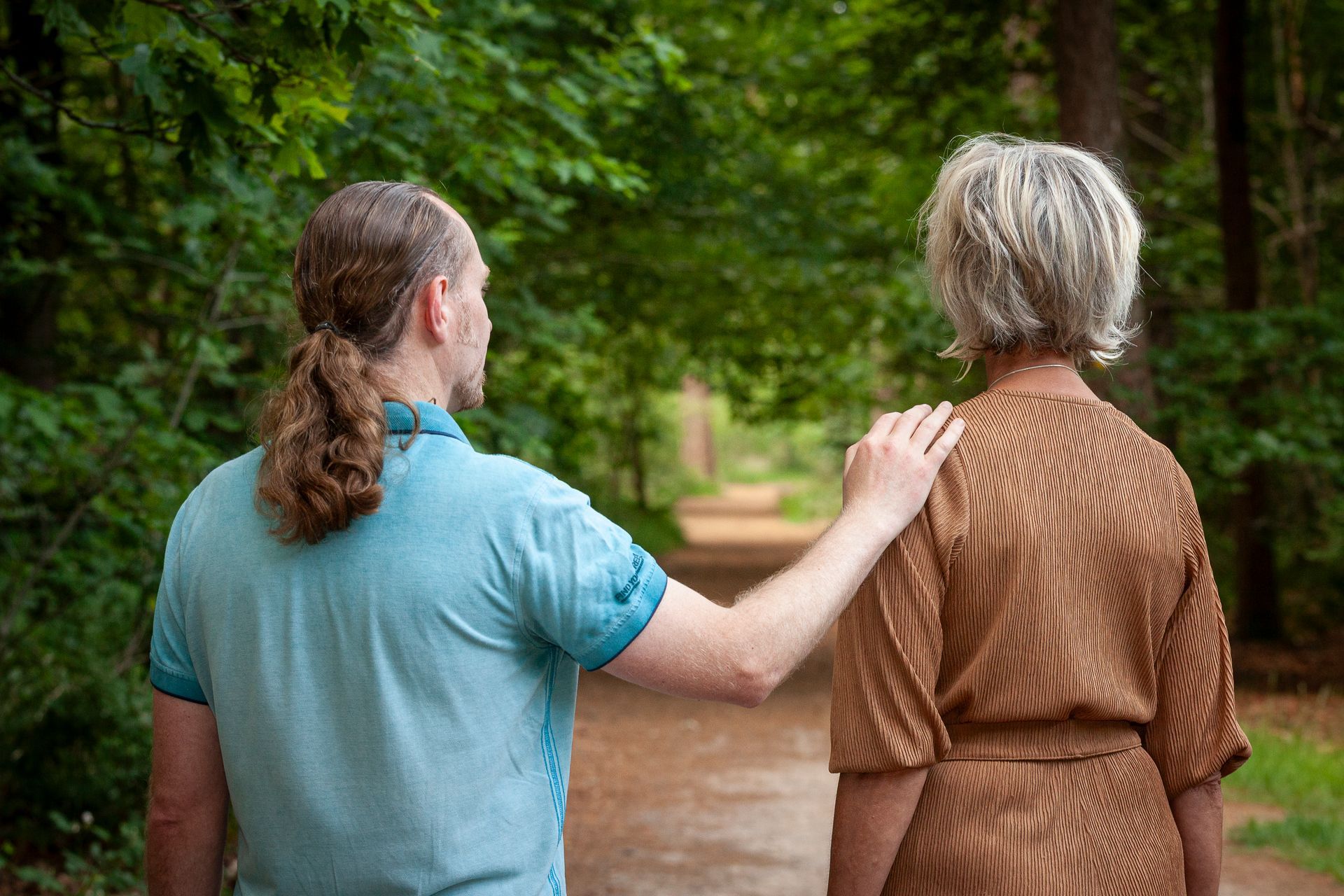 In een bos legt een persoon in een lichtblauw shirt een hand op de schouder van een andere persoon in een bruine jurk met patroon.