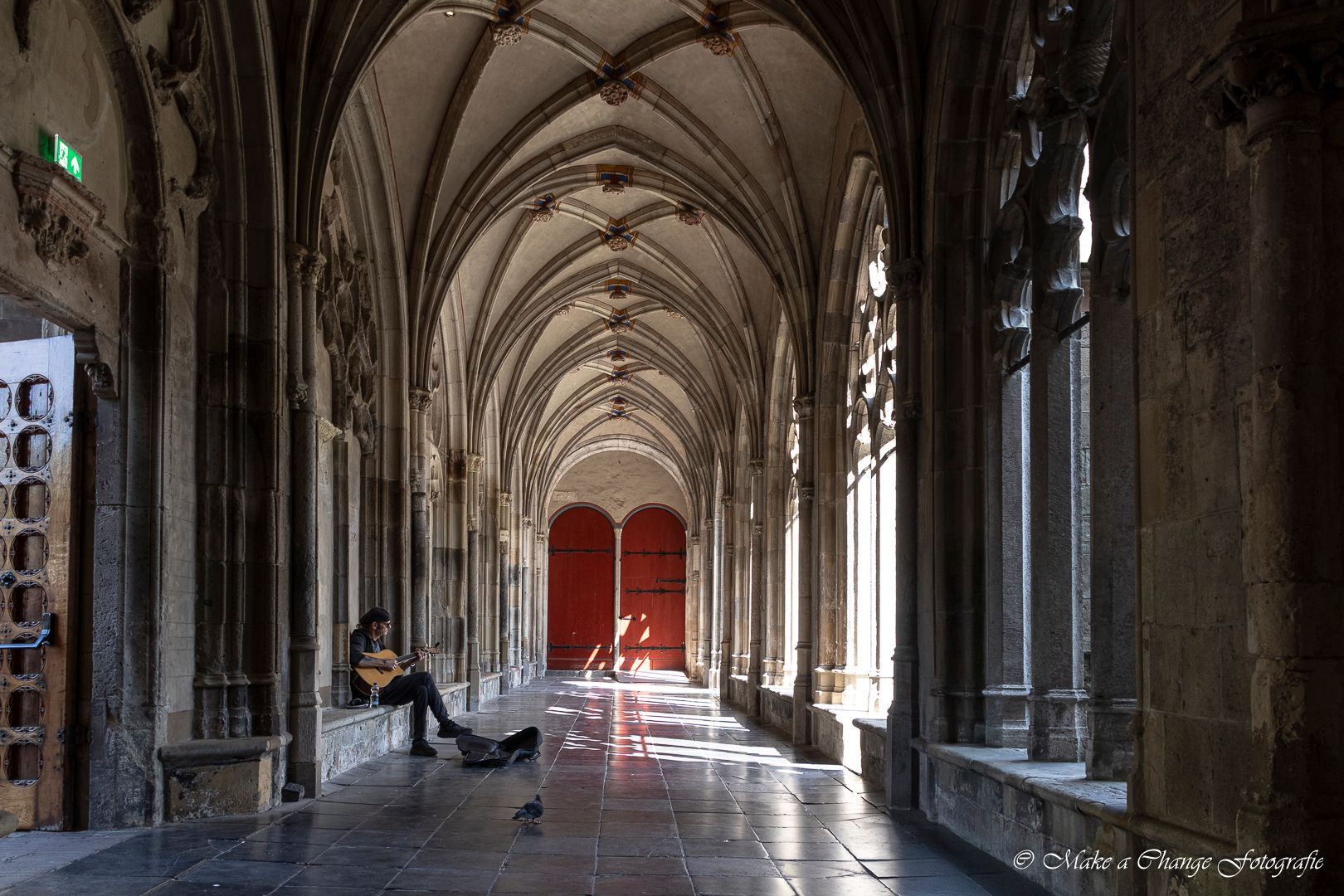 Een persoon speelt gitaar in een lange, stenen gang met gewelfde plafonds en zonlicht dat door de hoge ramen naar binnen stroomt.