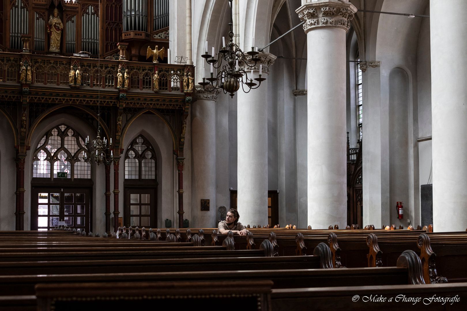 Interieur van een kerk met rijen houten banken, grote witte pilaren en sierlijke architectonische details. Iemand zit in de banken.