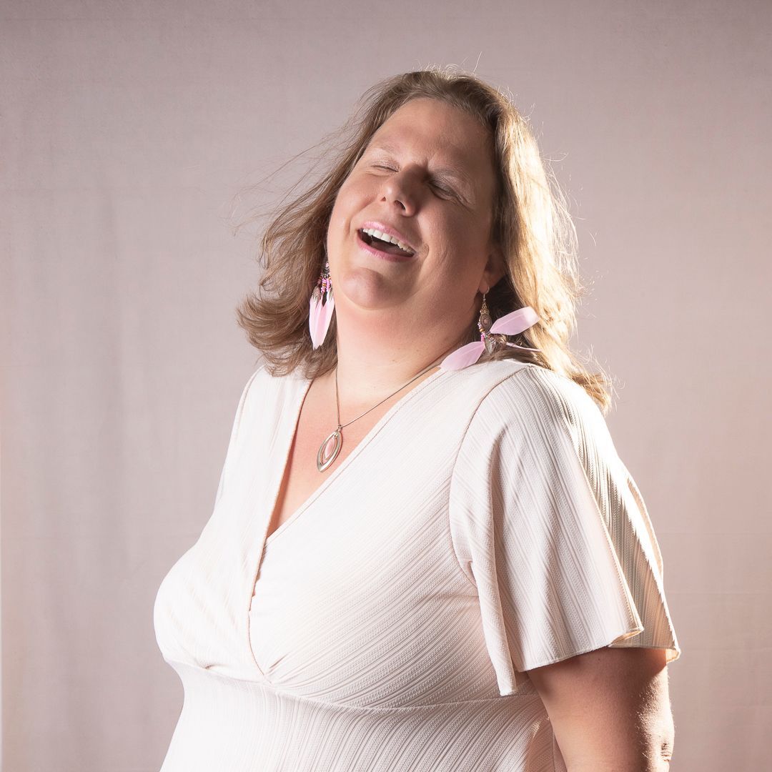 Een vrouw in een witte jurk lacht met haar hoofd naar achteren; ze draagt roze oorbellen en een ketting.