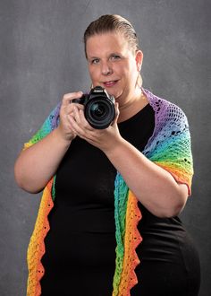 Vrouw met camera in de hand, gehuld in een regenboogkleurige sjaal, staand voor een grijze achtergrond.
