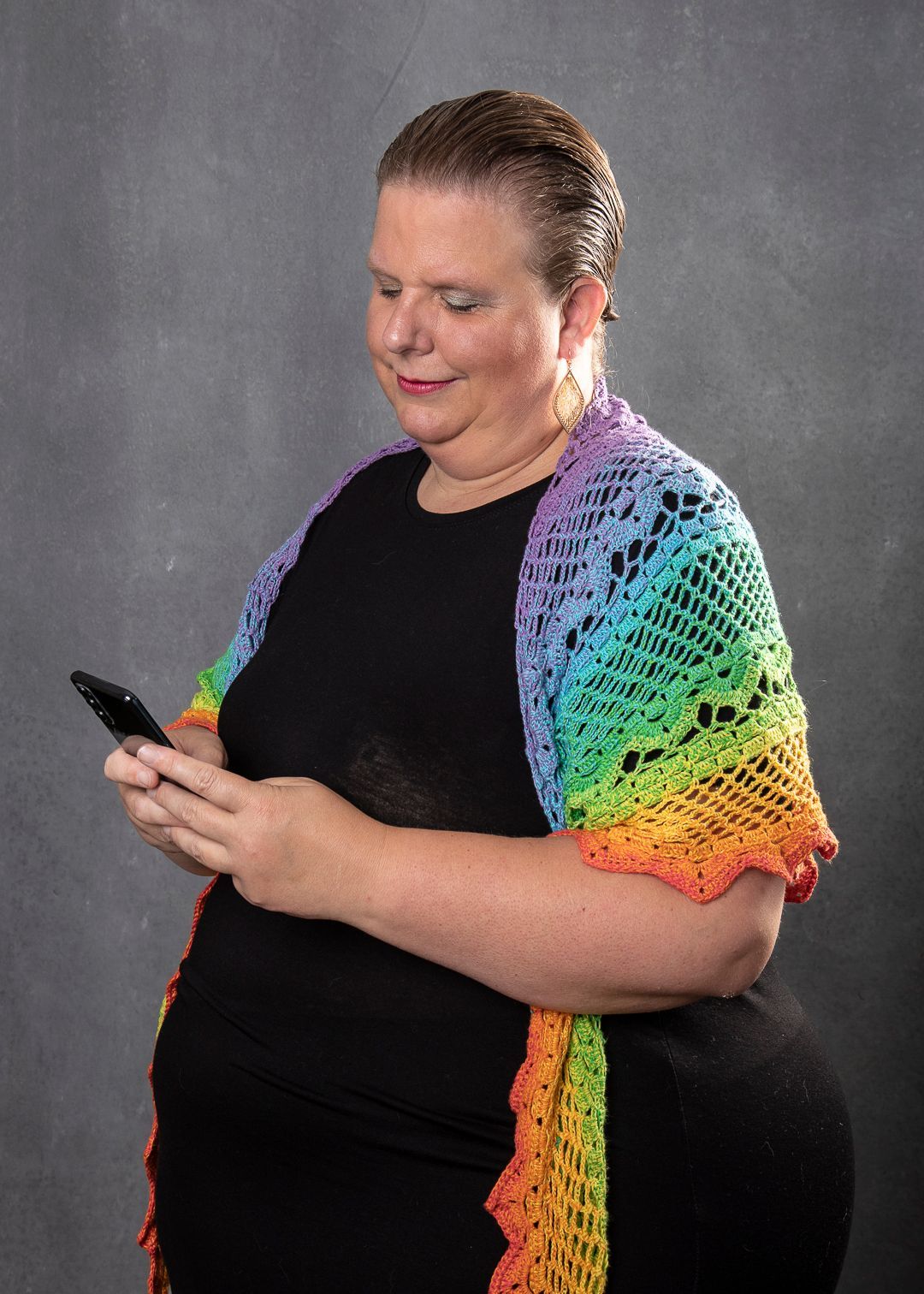 Vrouw in zwarte jurk, gehaakte regenboogsjaal, kijkt naar haar telefoon. Grijze achtergrond. Make a Change Call
