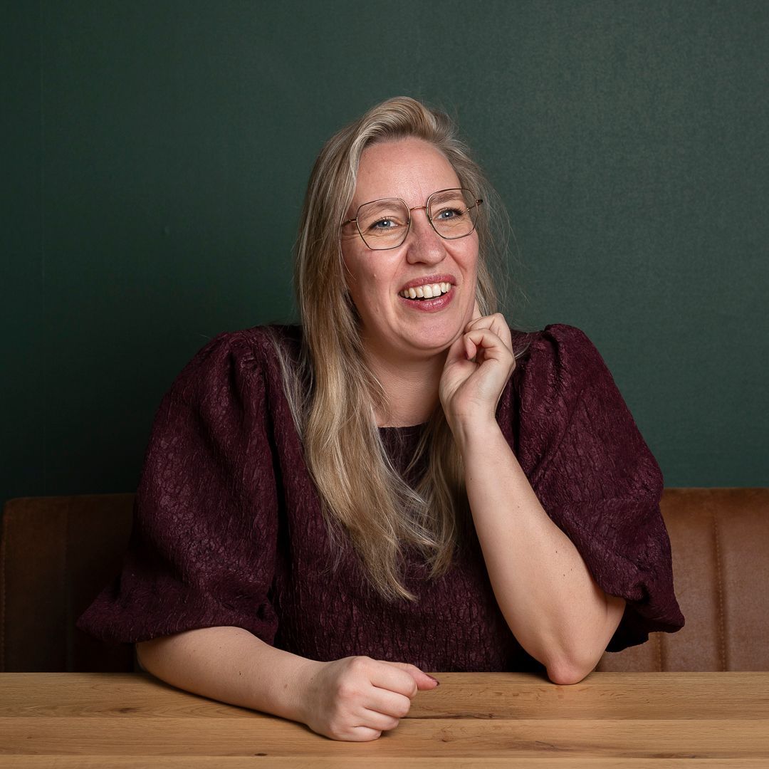 Een persoon met lang blond haar en een bril, gekleed in een paarse top met textuur, glimlacht en laat zijn kin op een hand rusten.