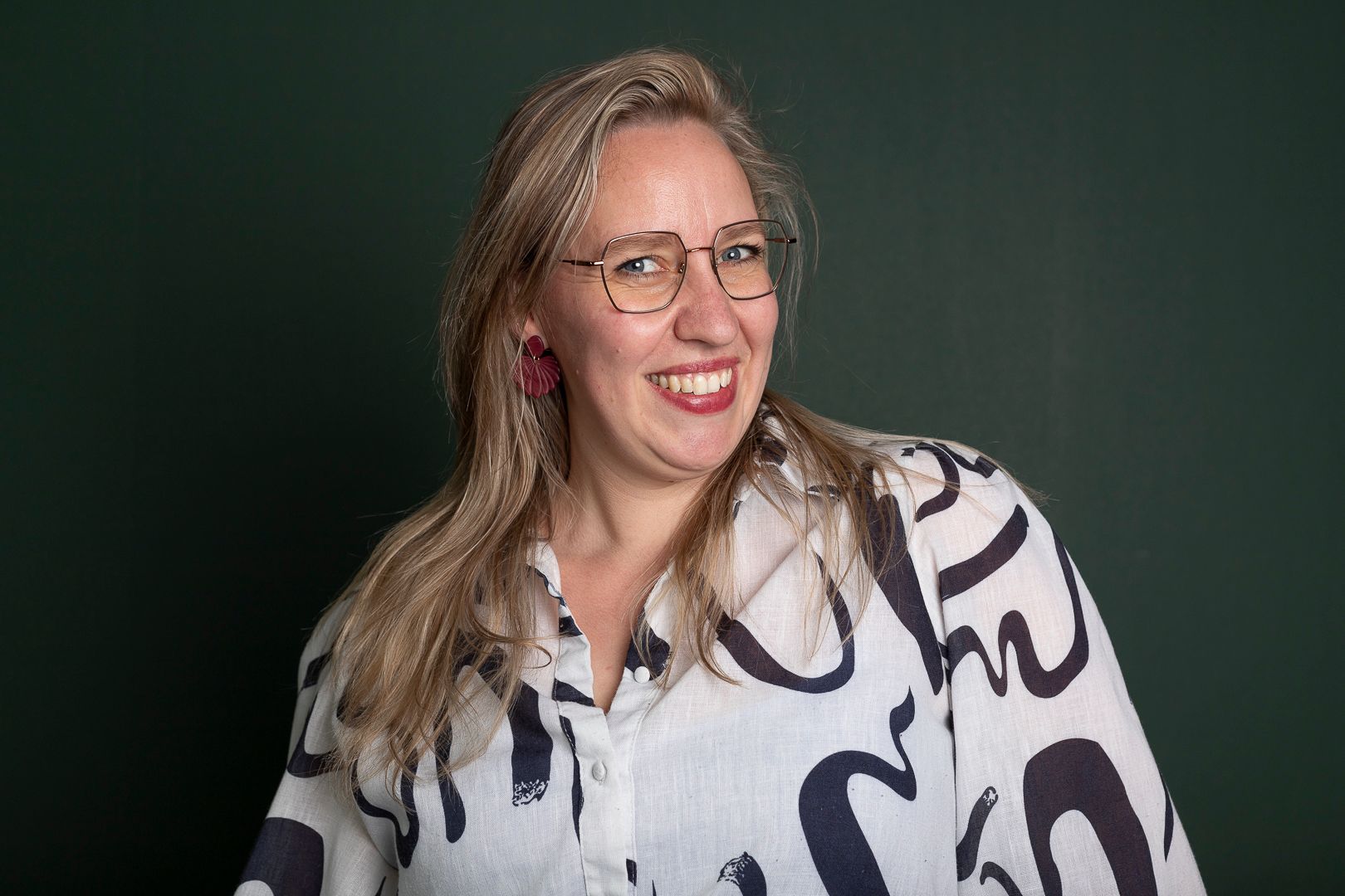 Een lachend persoon met een bril en een witte blouse met een abstract zwart patroon tegen een donkergroene achtergrond.