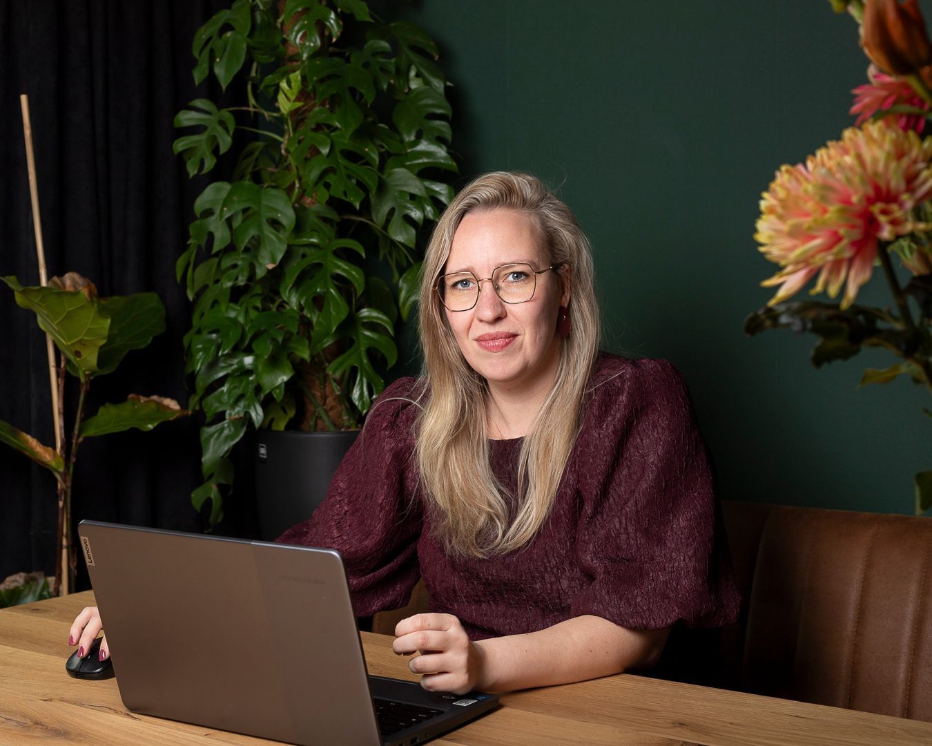Een persoon met een bril en een bordeauxrode top werkt op een laptop aan een houten tafel voor een groene muur met kamerplanten.