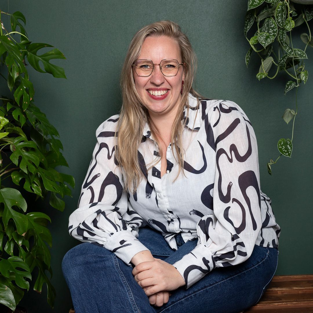 Een glimlachend persoon in een wit overhemd met patroon en een spijkerbroek zit tegen een donkergroene muur vol kamerplanten.