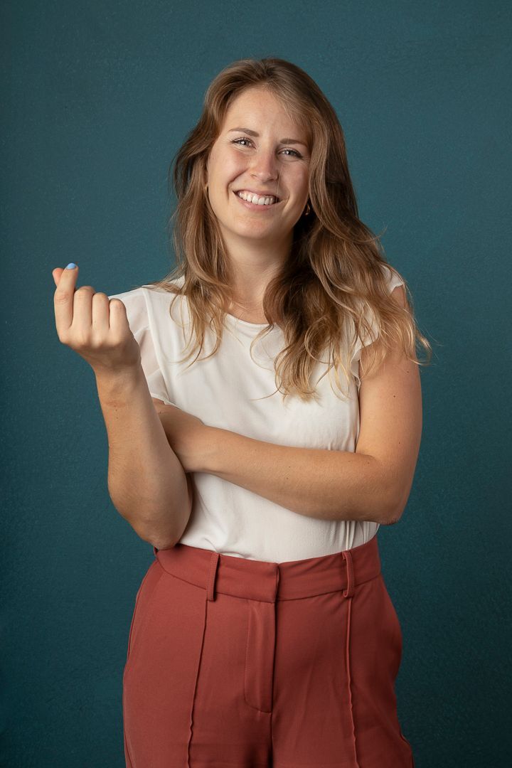 Een vrouw met bruin haar lacht en maakt een gebaar met één hand. Ze draagt een wit shirt en een roestkleurige broek op een blauwe achtergrond.