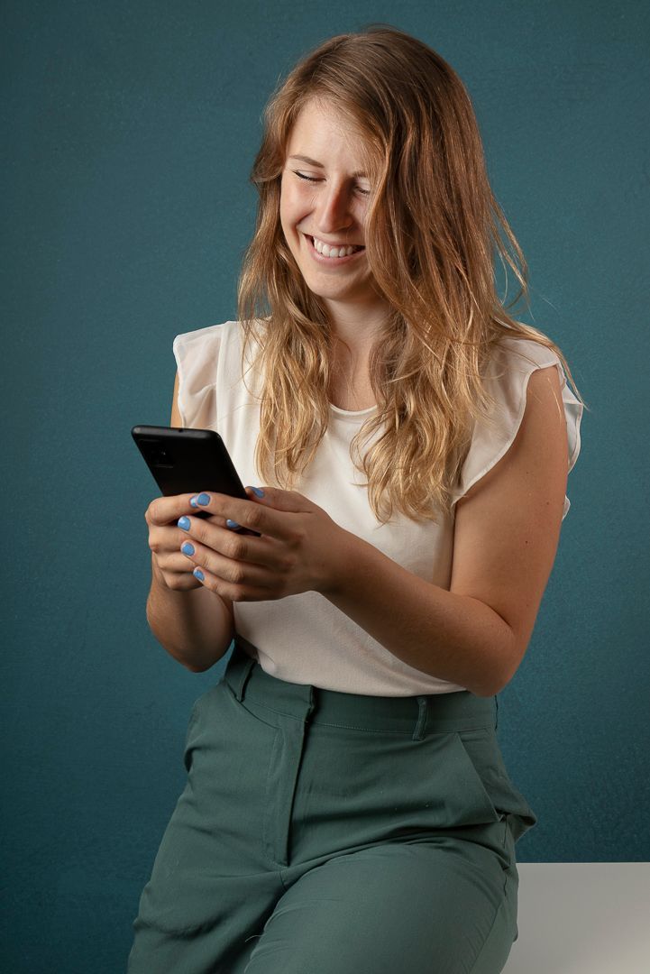 Vrouw met blond haar, gekleed in een witte blouse en een groene broek, lacht terwijl ze op haar telefoon kijkt tegen een blauwgroene achtergrond.