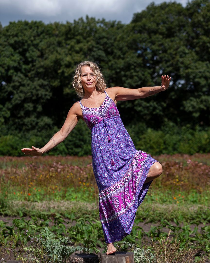Vrouw in een paarse jurk balanceert buiten op één been, armen uitgestrekt, lachend.