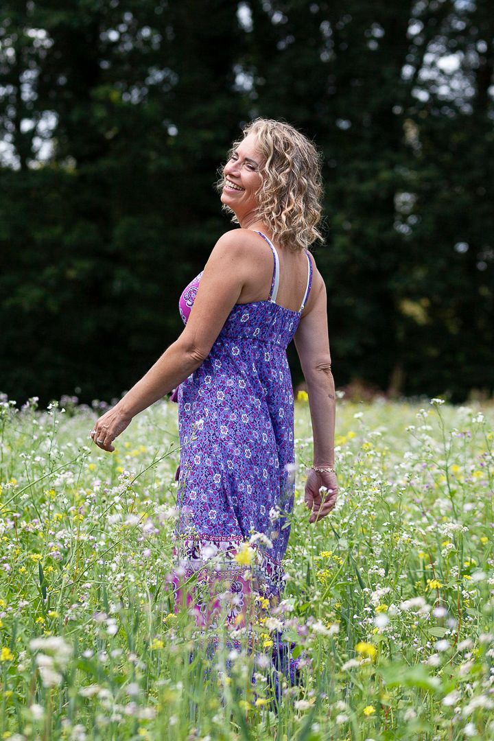Een vrouw met blonde krullen in een paarse bloemenjurk glimlacht terwijl ze door een veld vol wilde bloemen loopt.