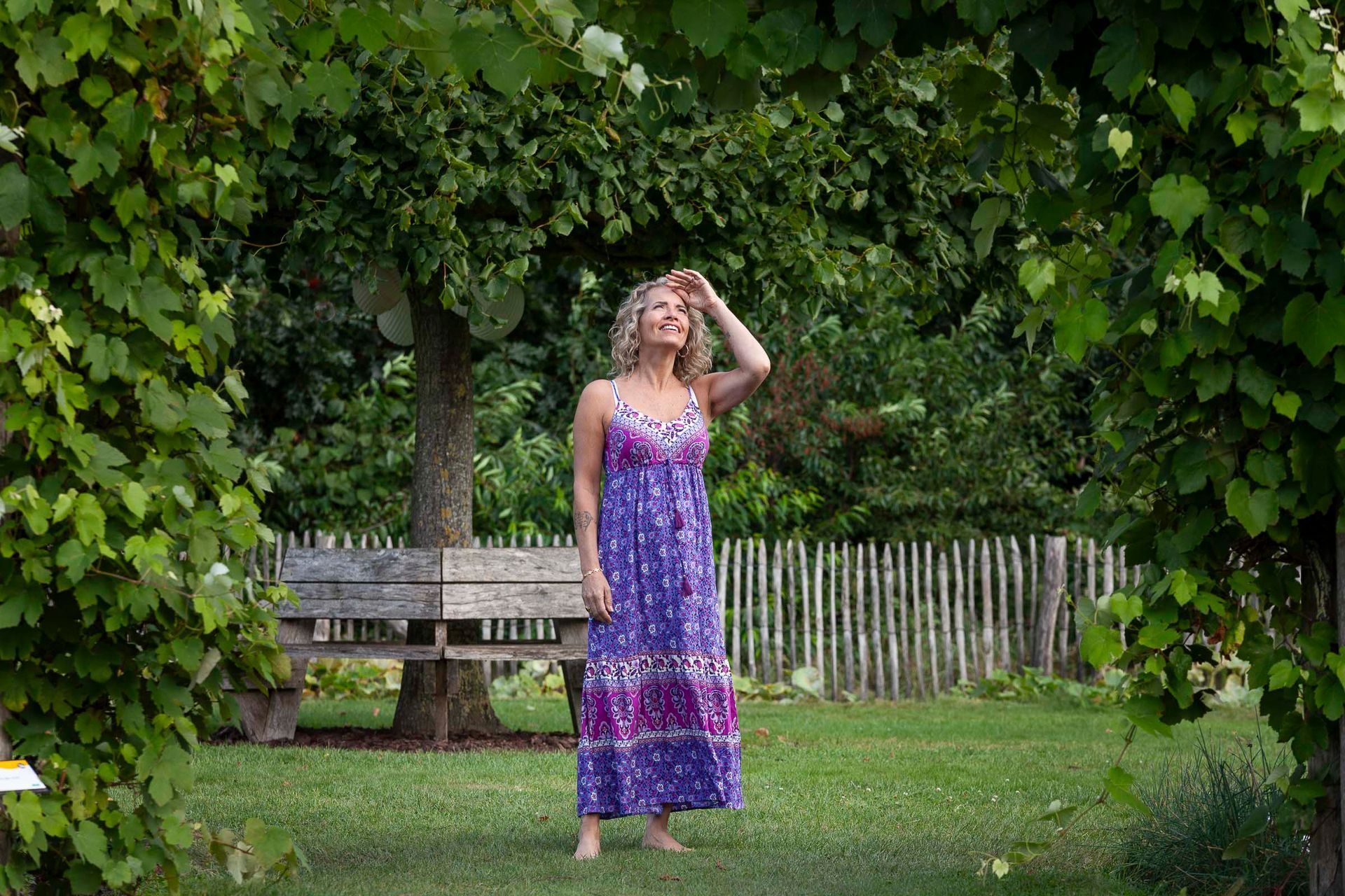 Een vrouw in een paarse jurk staat in een tuin, haar armen omhoog, kijkend naar de zon. Het tafereel is omlijst door wijnranken en er zijn een bankje en een hek zichtbaar.