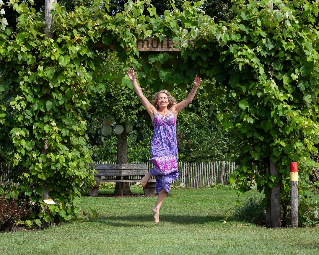 Een vrouw springt van vreugde onder een boog van druivenranken. Ze draagt een paarse jurk en lacht in een zonnige tuin.