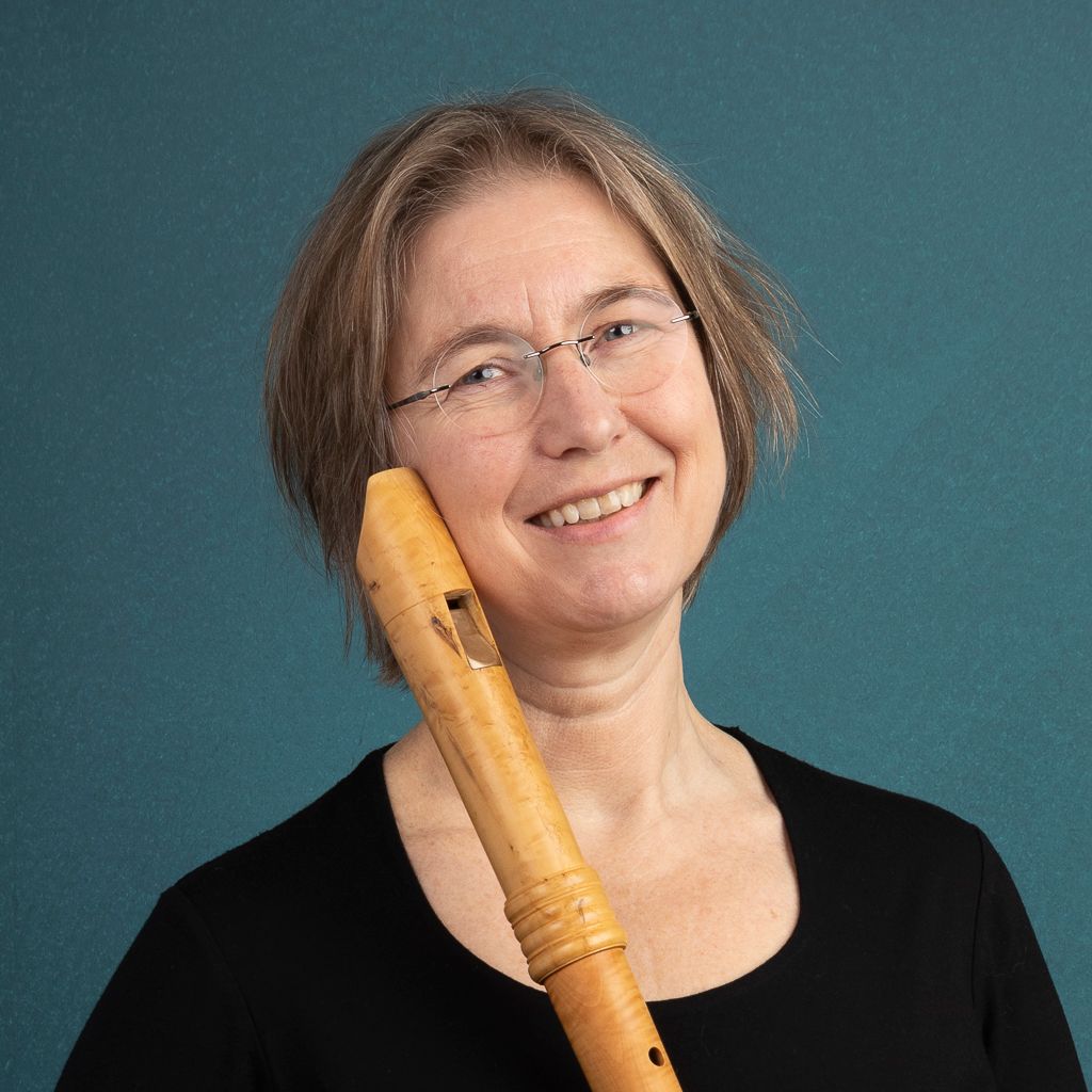 Lachende vrouw met bril en zwart shirt, met een recorder in haar hand. Blauwgroene achtergrond.