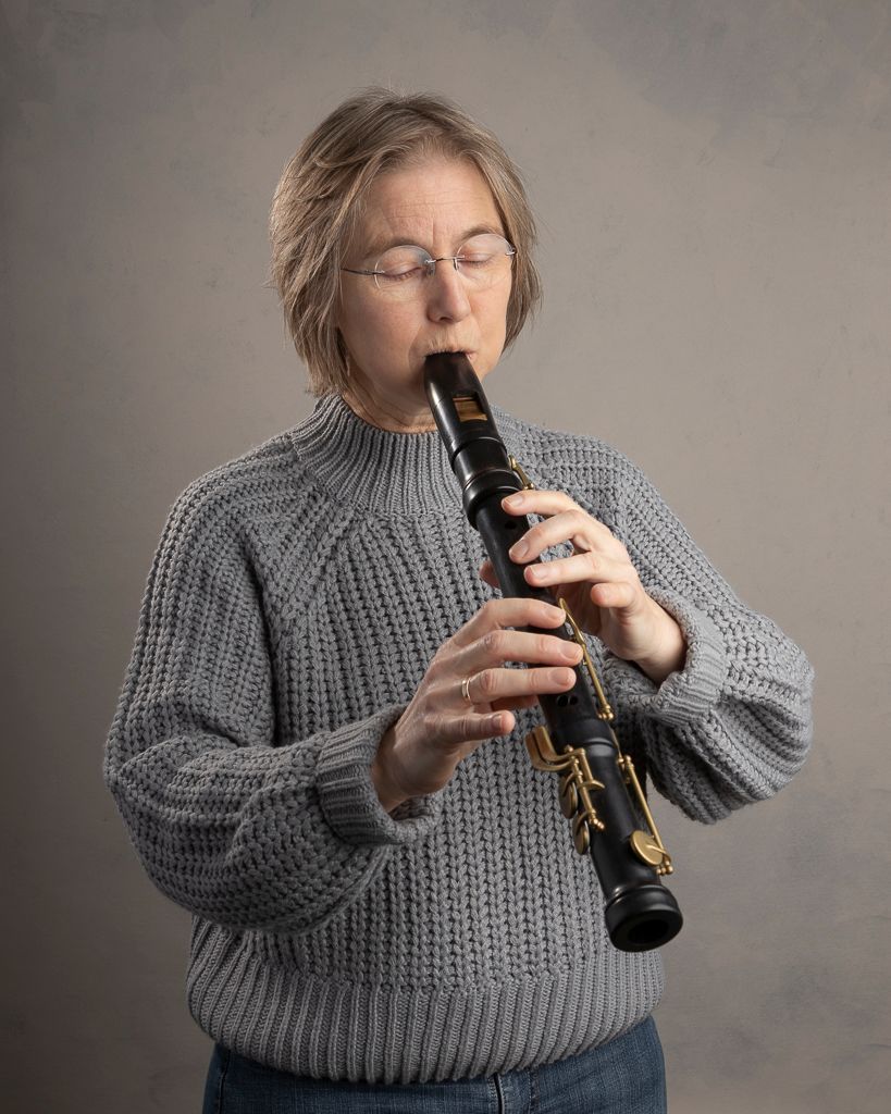 Een vrouw met een bril speelt op een zwarte klarinet, haar ogen gesloten, gekleed in een grijze trui. Ze staat tegen een grijze achtergrond.