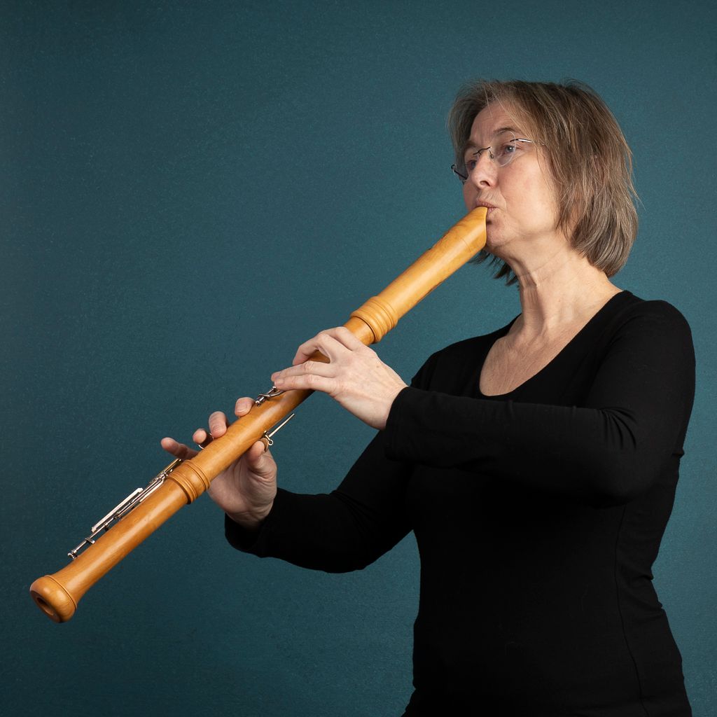 Een vrouw in het zwart speelt op een lange houten blokfluit tegen een blauwgroene achtergrond. Ze draagt een bril en heeft kort grijs haar.