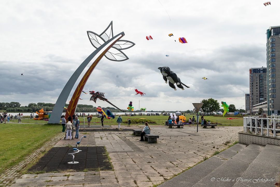 Vliegerfestival op een bewolkte dag aan een rivier met een modern kunstbeeld en verschillende kleurrijke vliegers in de lucht.